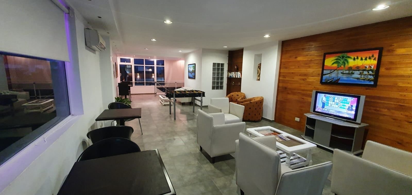 Sala de estar del hotel con sillones blancos, TV, mesitas y el metegol al fondo